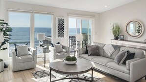 Een moderne woonkamer met een groot raam met uitzicht op zee, een grijze bank, een ronde salontafel en een klein bijzettafeltje met een lamp.