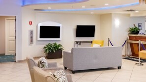 A hotel lobby with a reception desk, two flat-screen TVs mounted on the wall, a potted plant, a sofa with patterned cushions, and a chair.