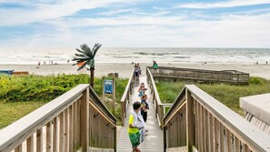 A wooden boardwalk leading to a beach with a palm tree and a sign.
