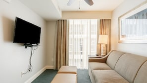 A hotel room with a flat-screen TV mounted on the wall, a sofa, an ottoman, a lamp on a side table, and a window with curtains. - North Myrtle Beach | Family Wave Hub | Pool + Beach | 3BR | Sleeps 10 (North Myrtle Beach)