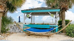 A hammock on a sandy beach, a gazebo with a blue roof, and palm trees.