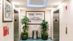 Un hall d’hôtel avec deux ascenseurs, des plantes en pot, un tableau encadré et une poubelle.