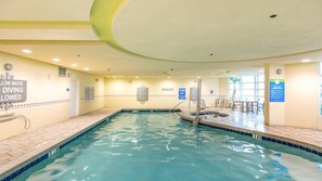 Ein Hallenbad mit klarem Wasser, Sprungbrett und Liegebereich mit Stühlen und Tischen.
