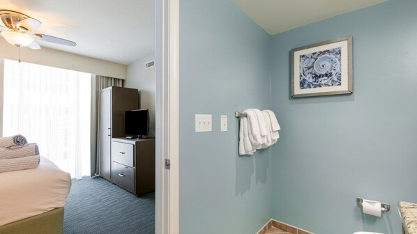 A hotel room with a bed, a desk, a television, a framed artwork, and a bathroom with a toilet and towel rack.