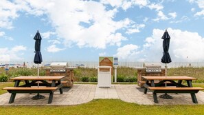 An outdoor patio area with two barbecue grills, picnic tables, and umbrellas. - North Myrtle Beach | Family Wave Hub | Pool + Beach | 3BR | Sleeps 10 (North Myrtle Beach)