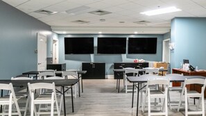 A conference room with tables, chairs, a large screen, and a desk with a computer. - North Myrtle Beach | Family Wave Hub | Pool + Beach | 3BR | Sleeps 10 (North Myrtle Beach)