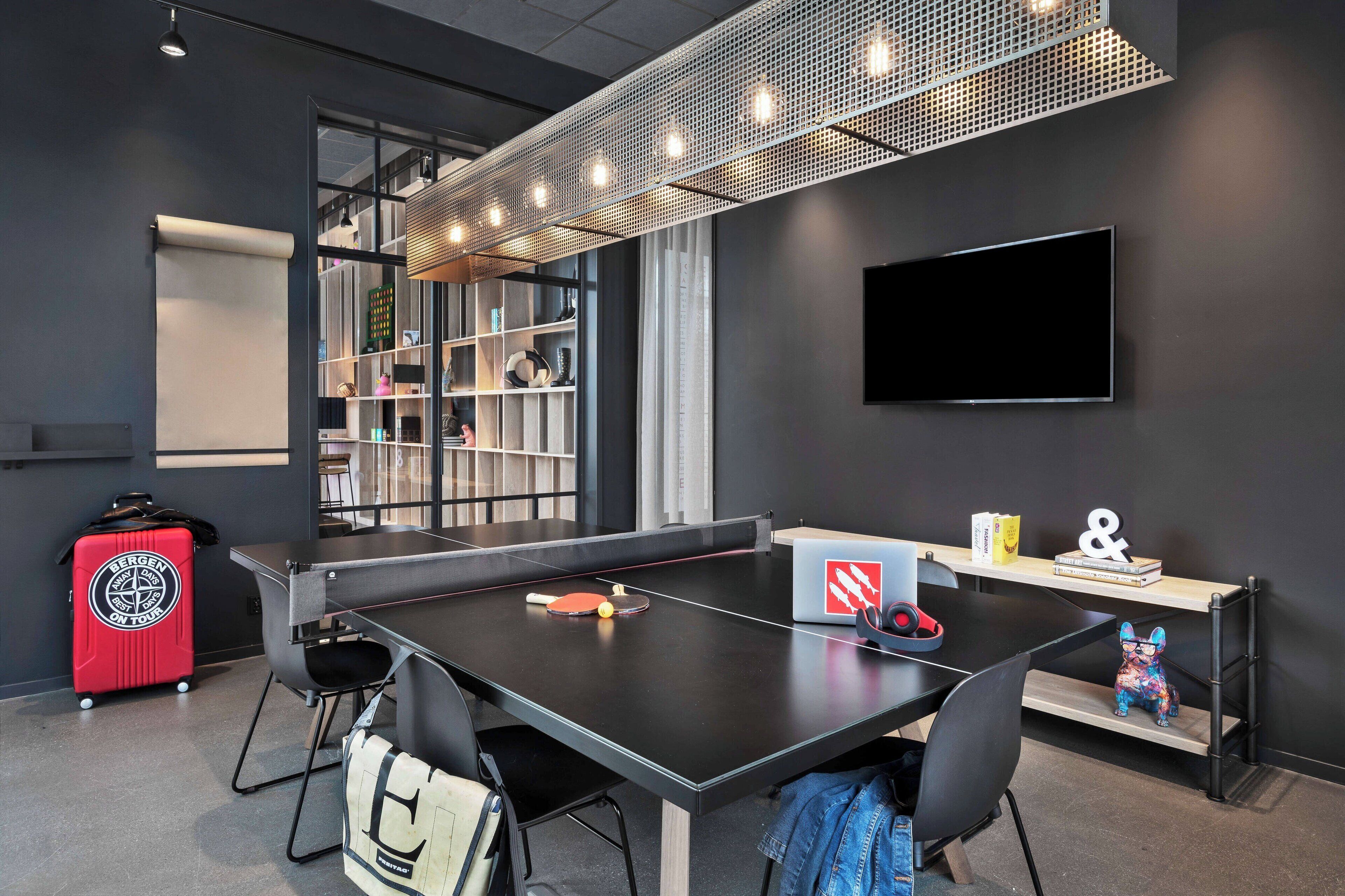 Una sala de reuniones moderna con una mesa de ping pong, sillas, una estantería, un televisor y una maleta roja.