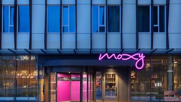 A modern building with a glass facade, a neon sign reading "Moxy," and a pink entrance area.