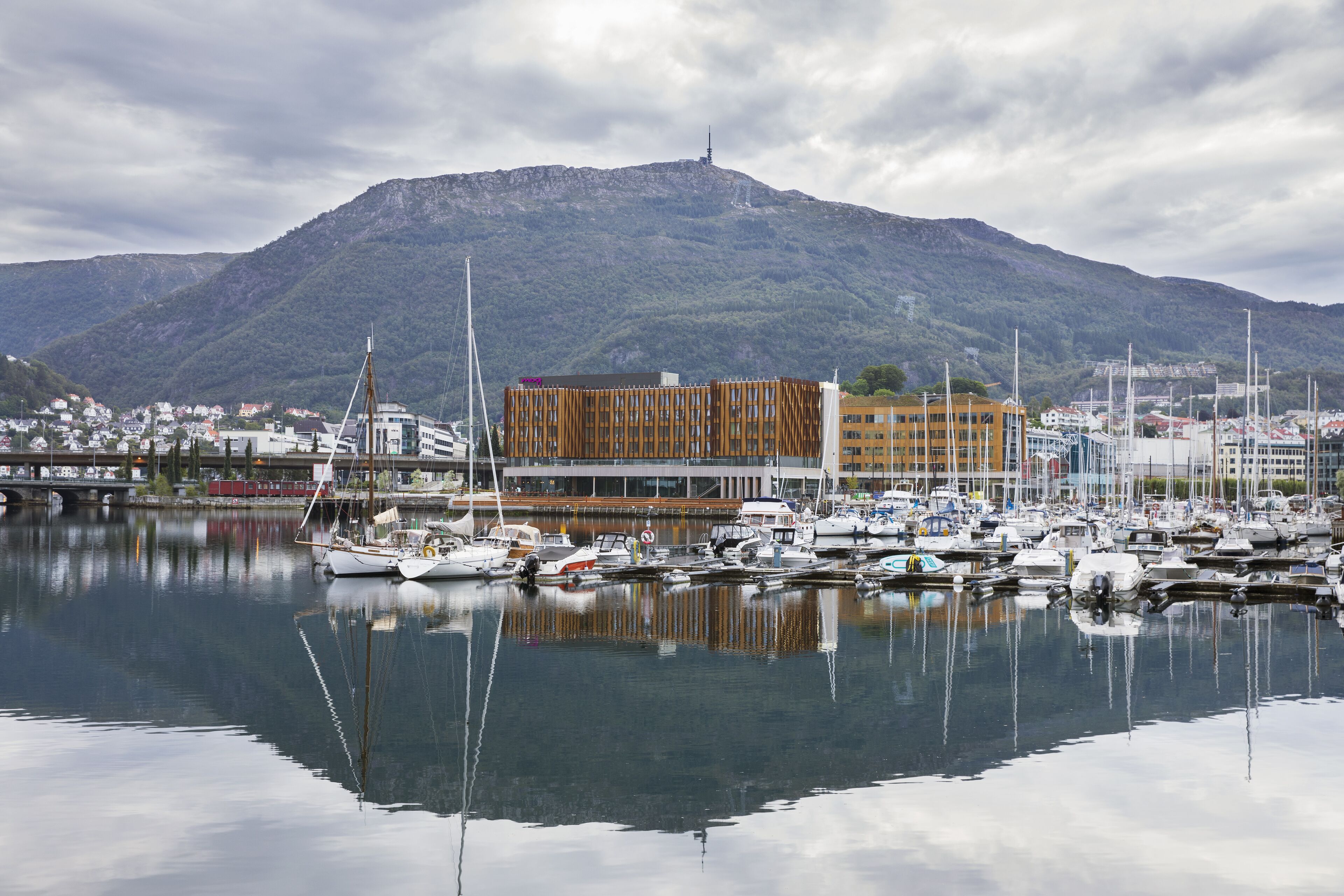 A marina with boats docked, a mountain in the background, and a bridge connecting to a town.