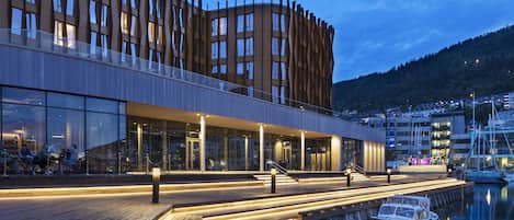 Un hotel moderno frente al agua, con muelle de madera, fachada de vidrio y vistas al agua y a las embarcaciones.