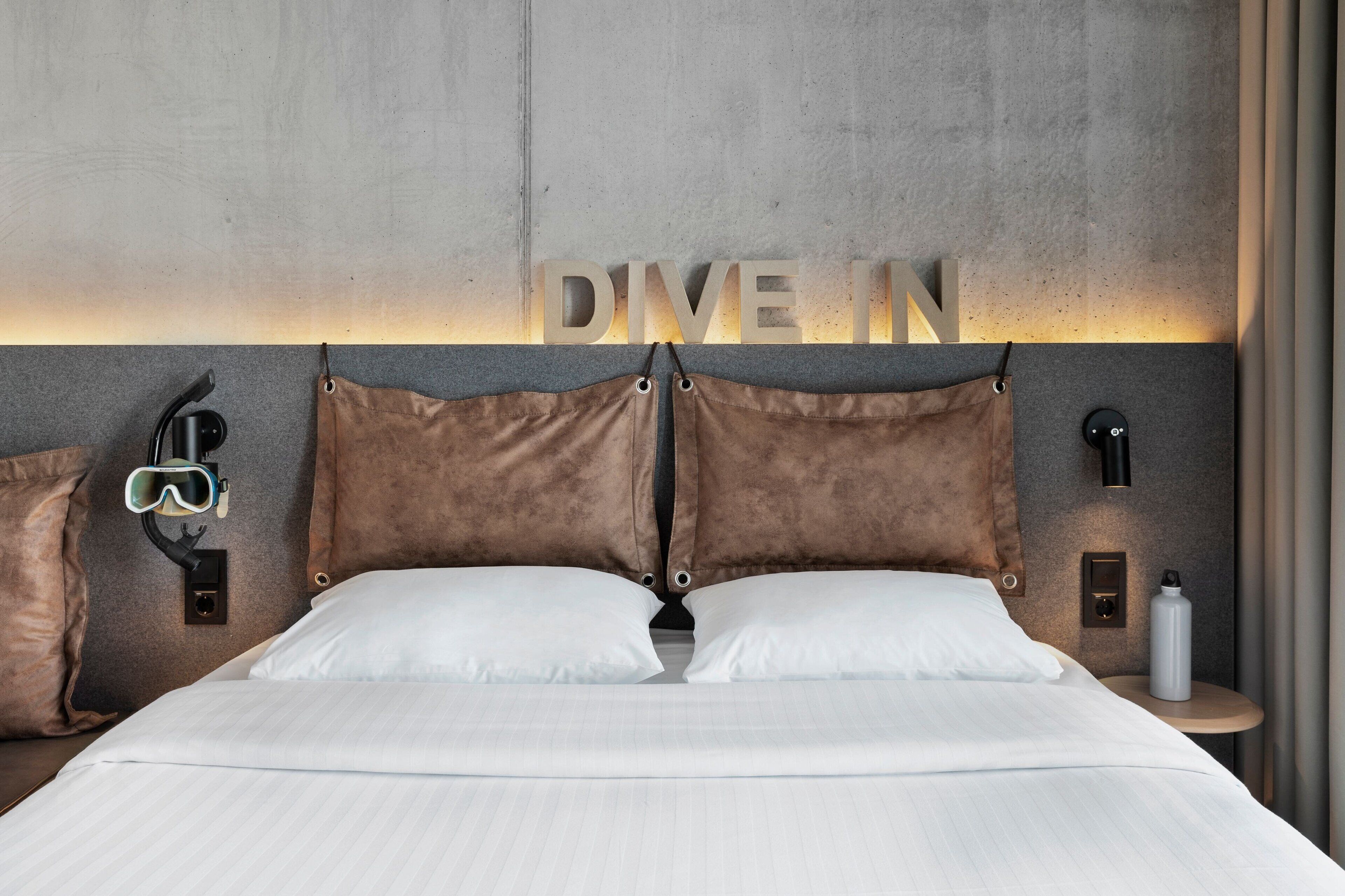 Ein modernes Hotelzimmer mit Bett, Kissen, einem Kopfteil mit leuchtenden 'DIVE IN'-Lettern, einer Wandleuchte, einem Nachttisch mit einer Flasche und einer Schutzbrille.