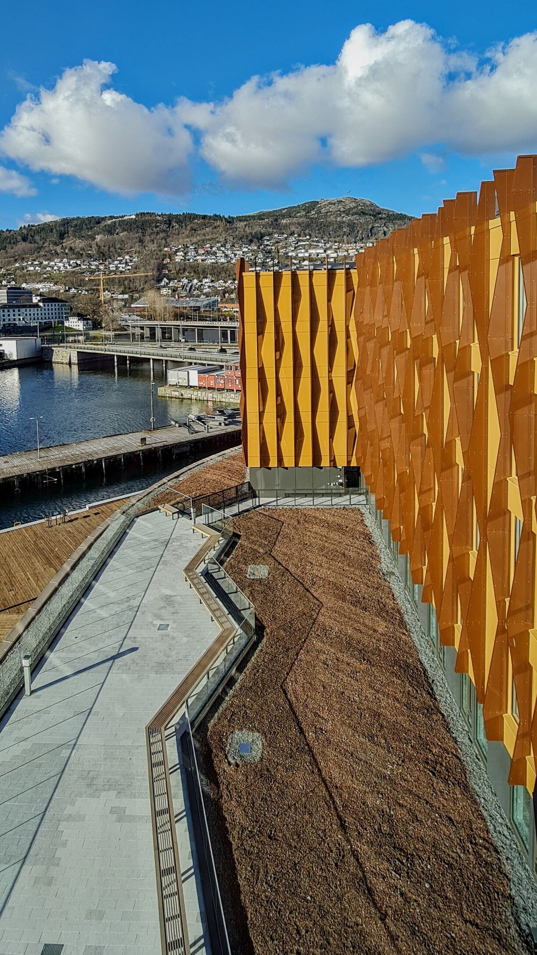 A waterfront building with a distinctive yellow and brown facade, a paved walkway, and a view of a residential area in the background.