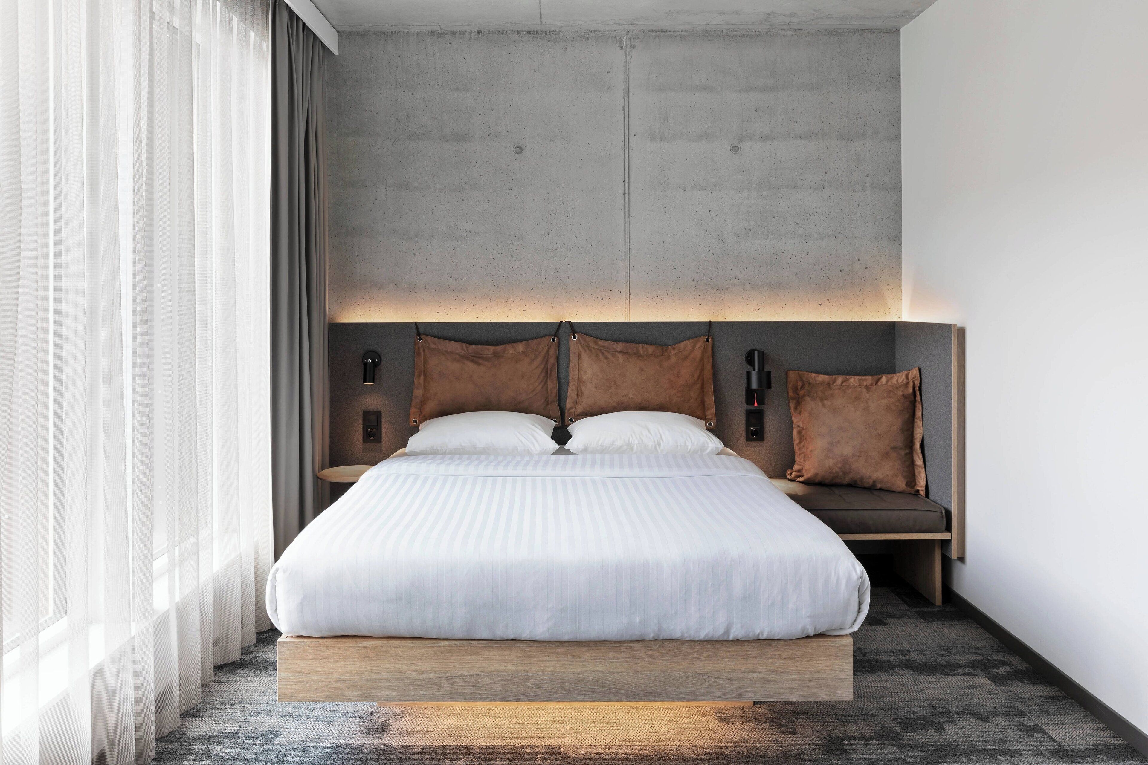 A modern hotel room with a large bed, a wooden platform, and a concrete headboard.