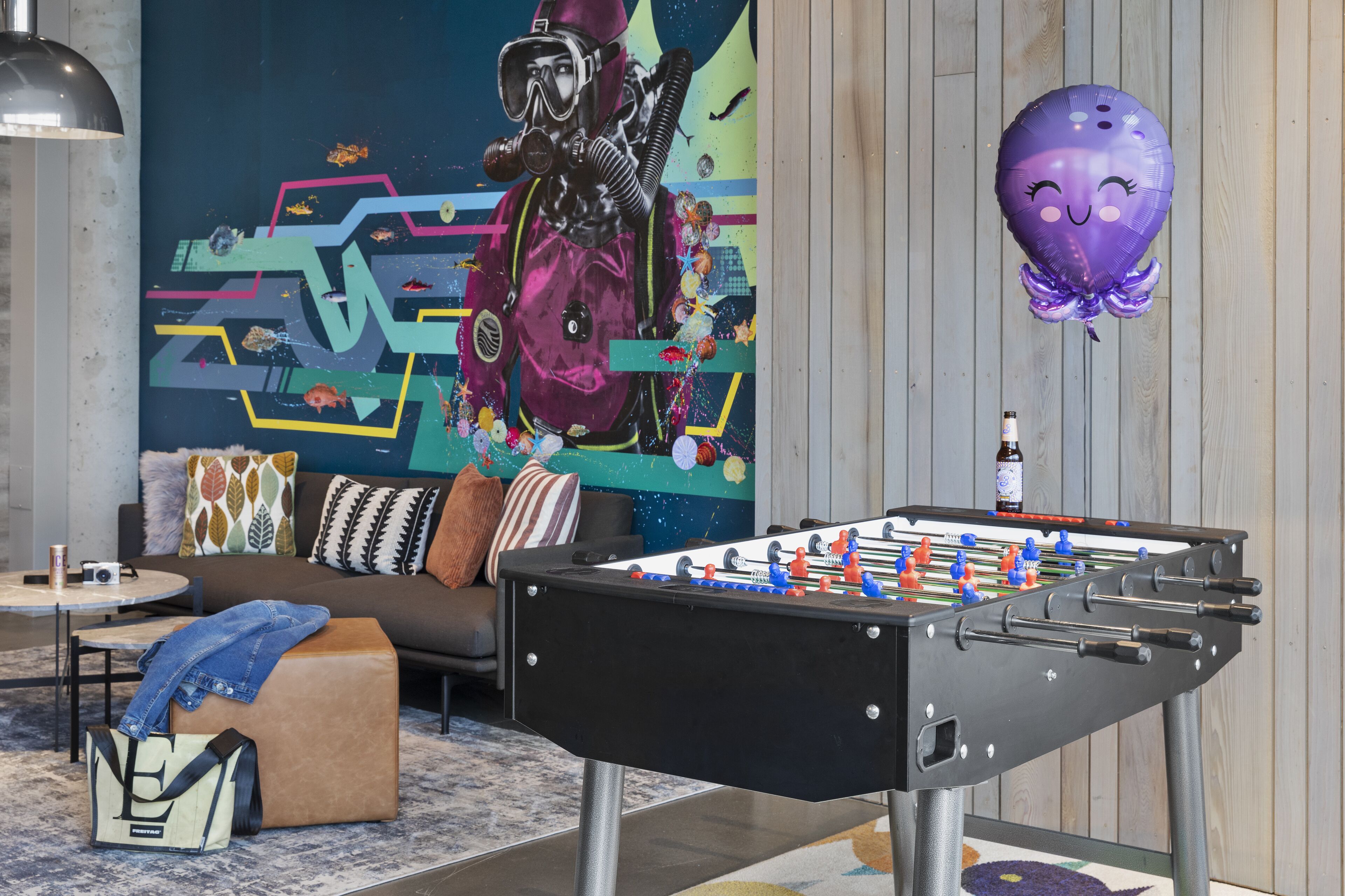 Una habitación con mesa de fútbol, sofá, mesa de centro y pared decorada con un mural colorido.