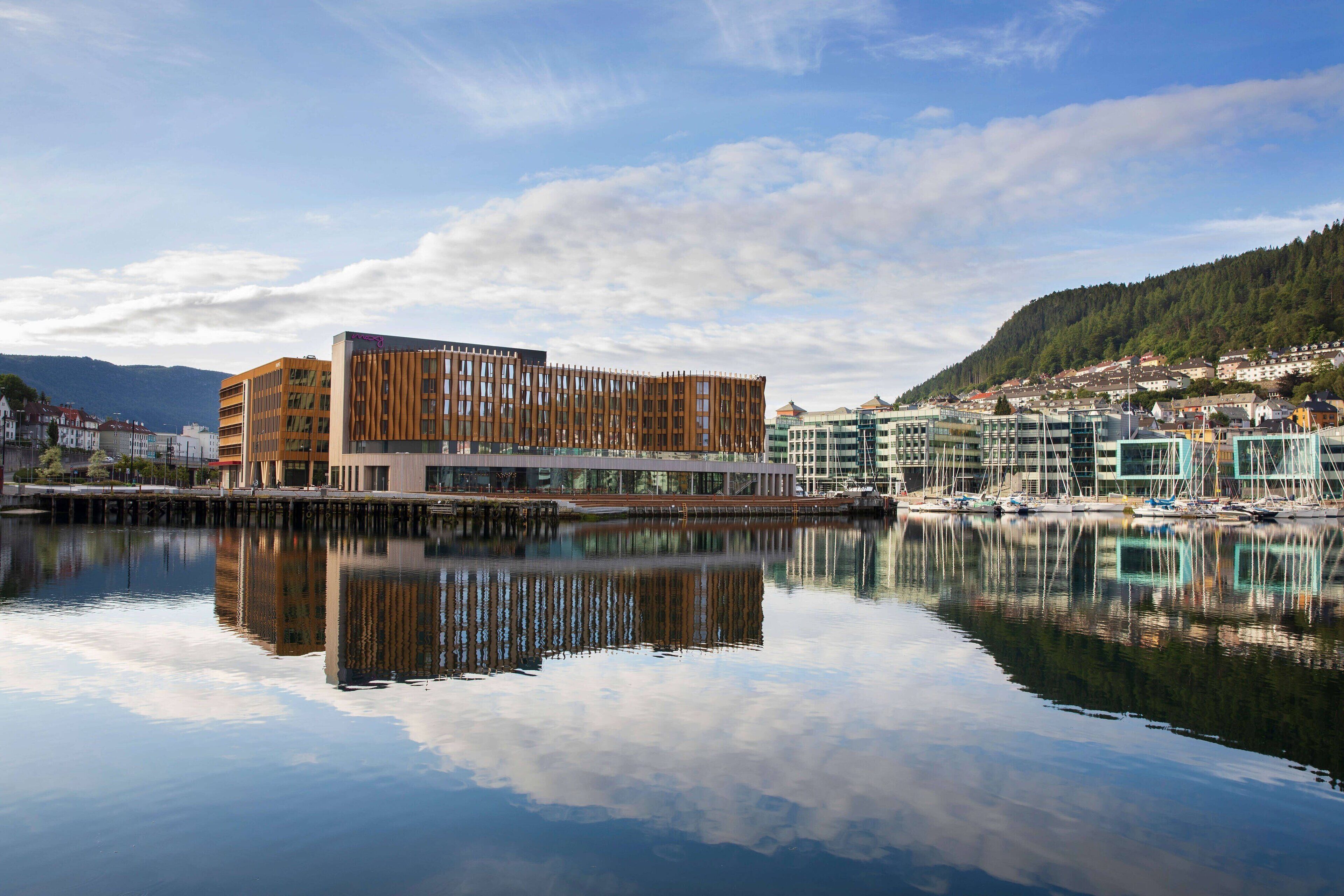 Un bâtiment situé au bord de l’eau, doté d’une architecture moderne, dont le reflet se mire dans l’eau calme, et qui est entouré d’autres bâtiments et d’un paysage vallonné.