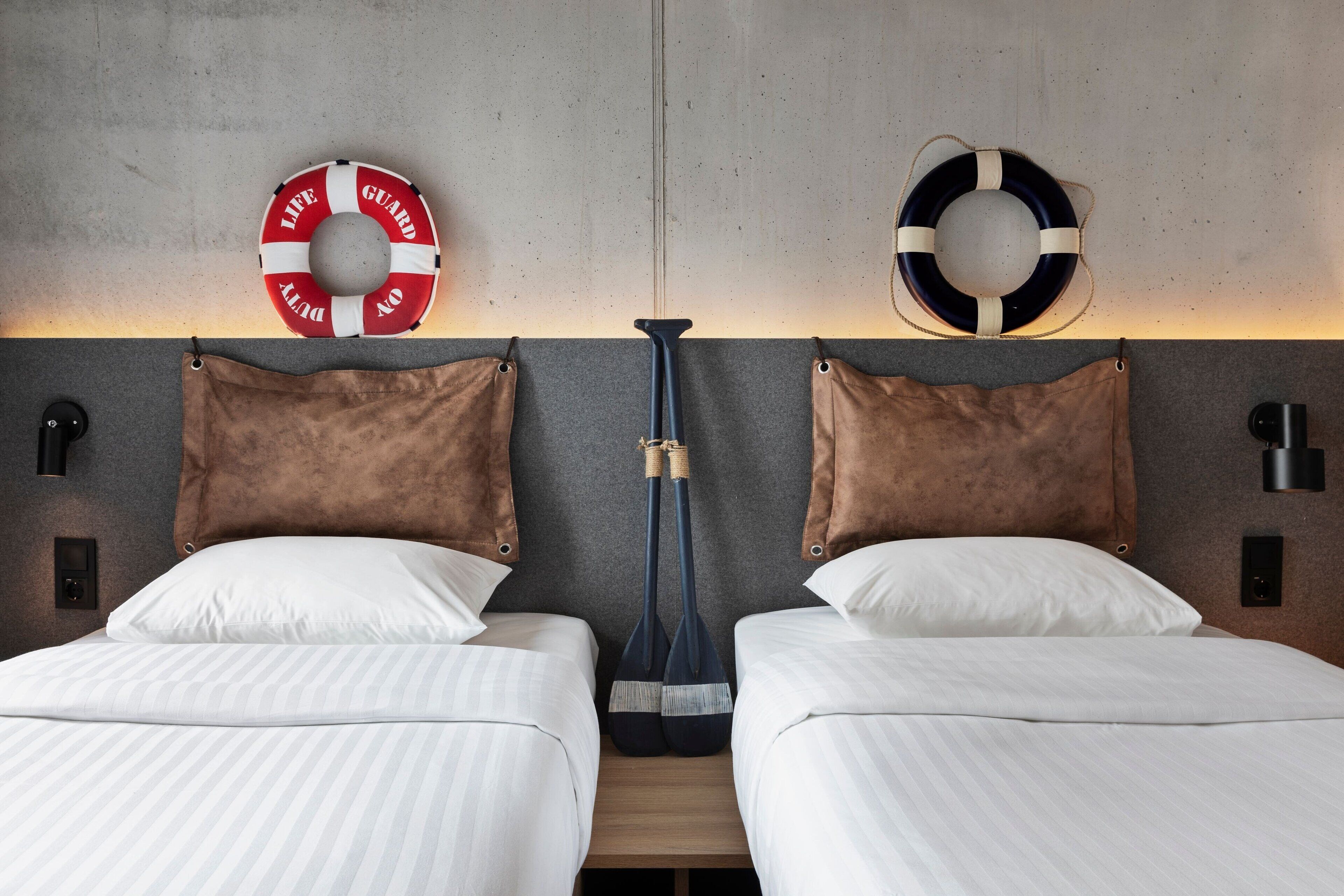 Ein Hotelzimmer mit zwei Betten, jeweils mit weißer Bettwäsche und braunen Kopfteilen. An der Wand über den Betten hängen Rettungsringe.