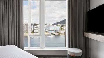 A hotel room with a large window, a bed, a TV, and a small stool.