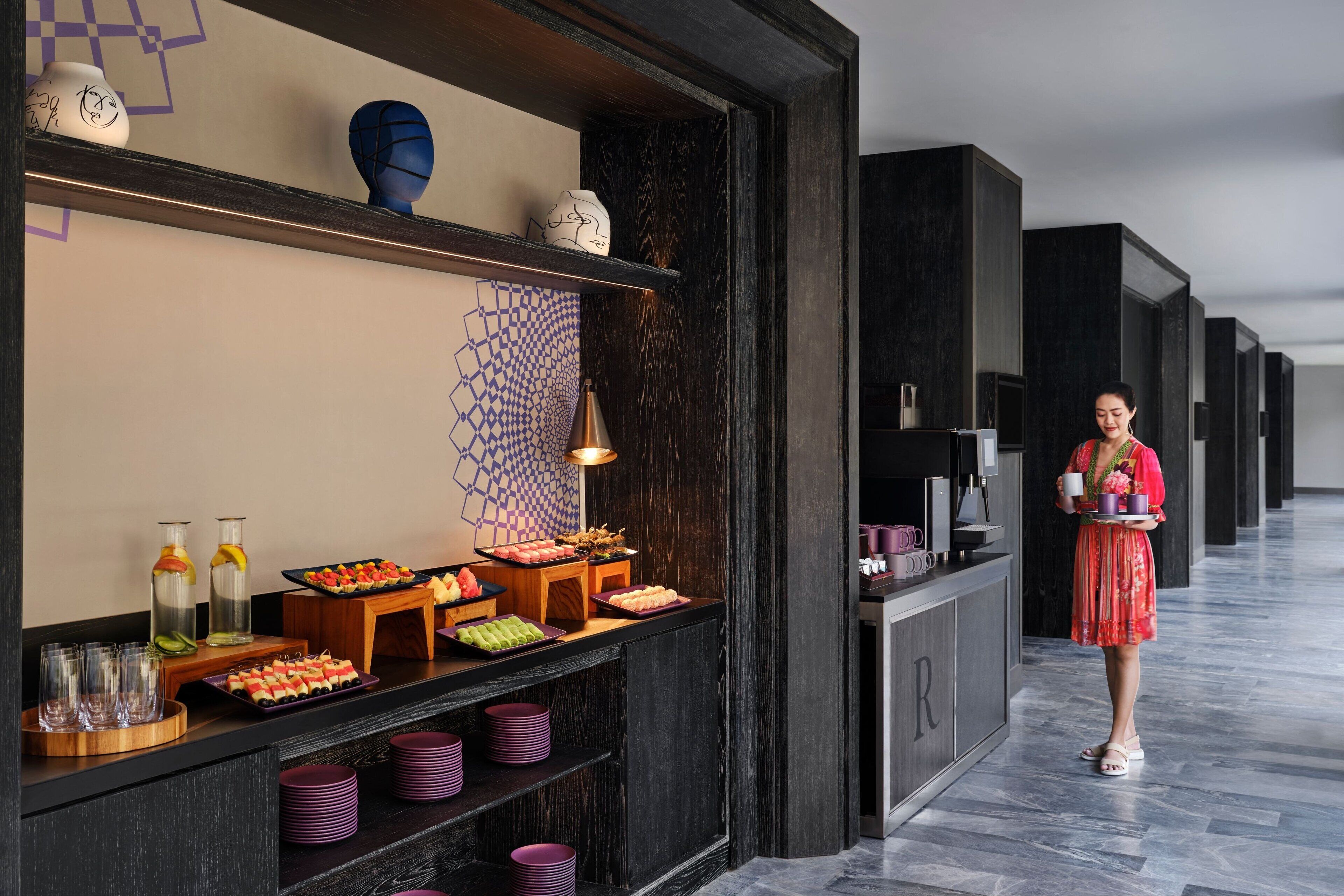 A hotel reception area with a buffet counter, a woman holding a tray, and a modern interior design.