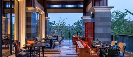 A spacious outdoor seating area with tables and chairs, a wooden bench with cushions, and a view of greenery in the background.