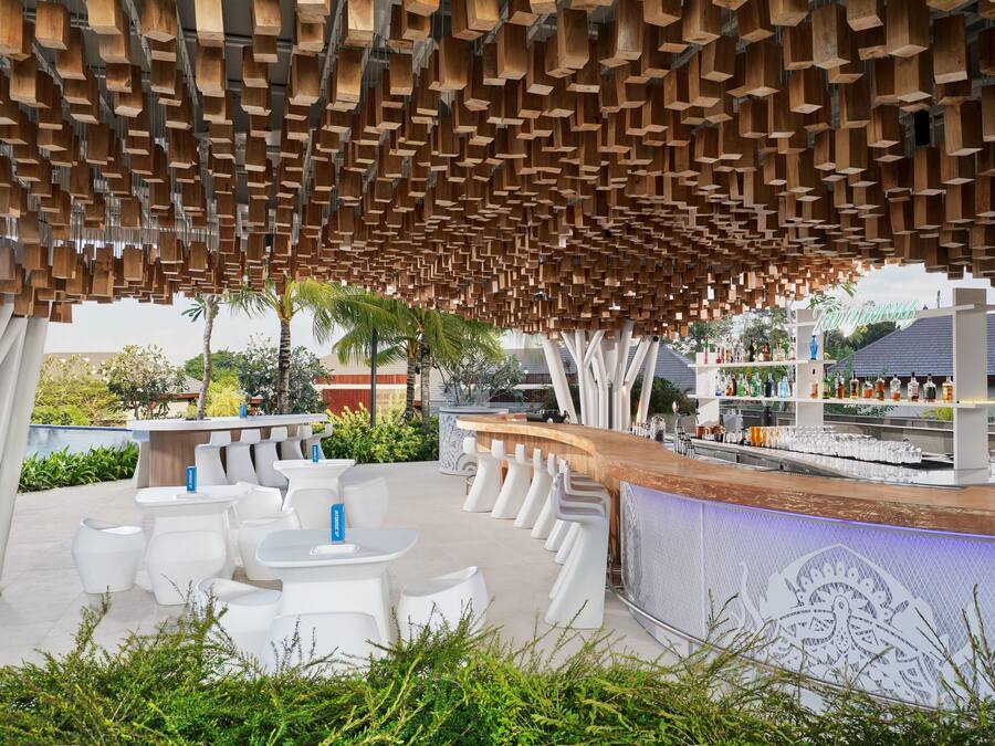 Een terras met witte stoelen en tafels, een houten balkenplafond en een bar met flessen.
