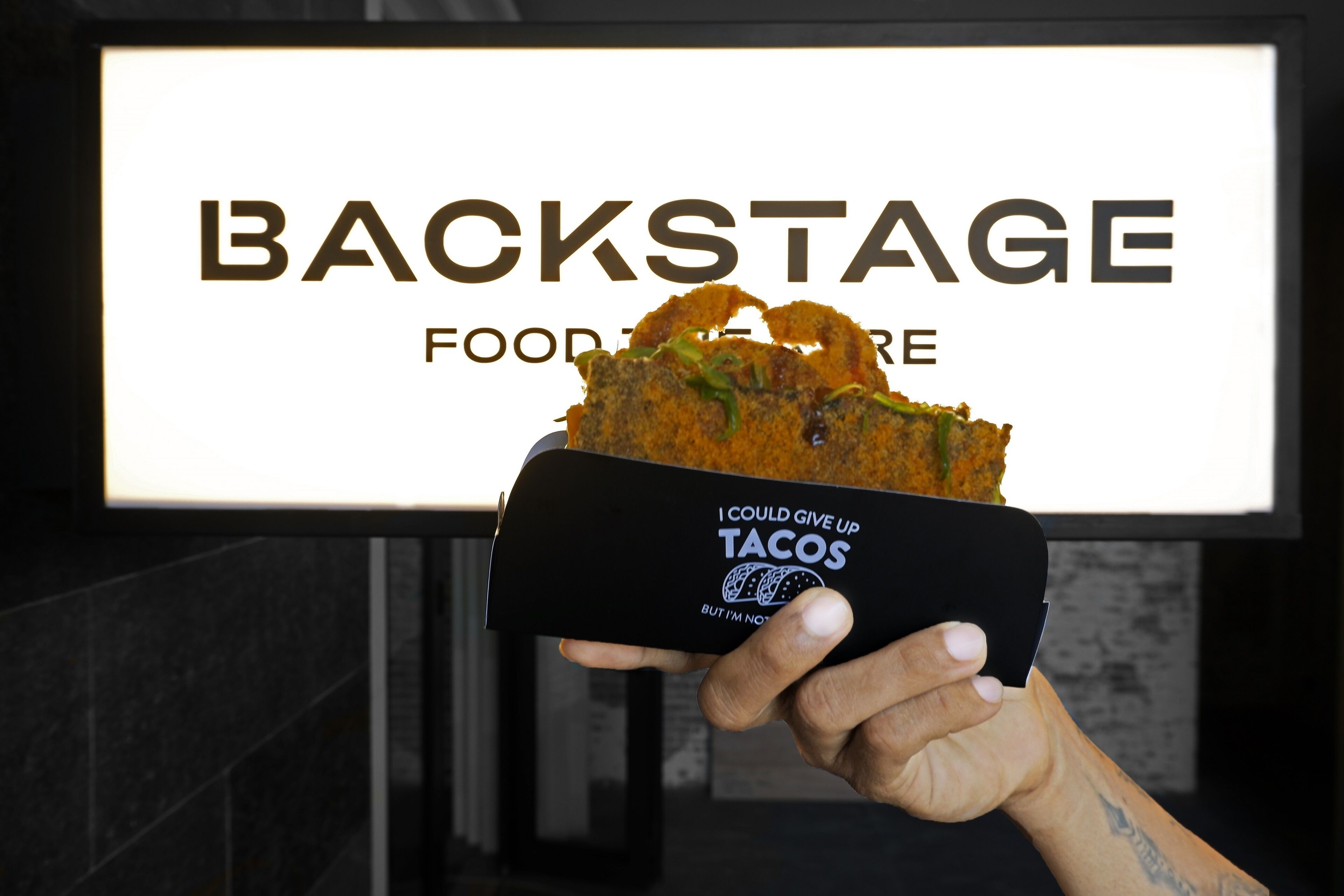 A hand holding a box of tacos with a sign in the background that reads 'BACKSTAGE FOODRE'.