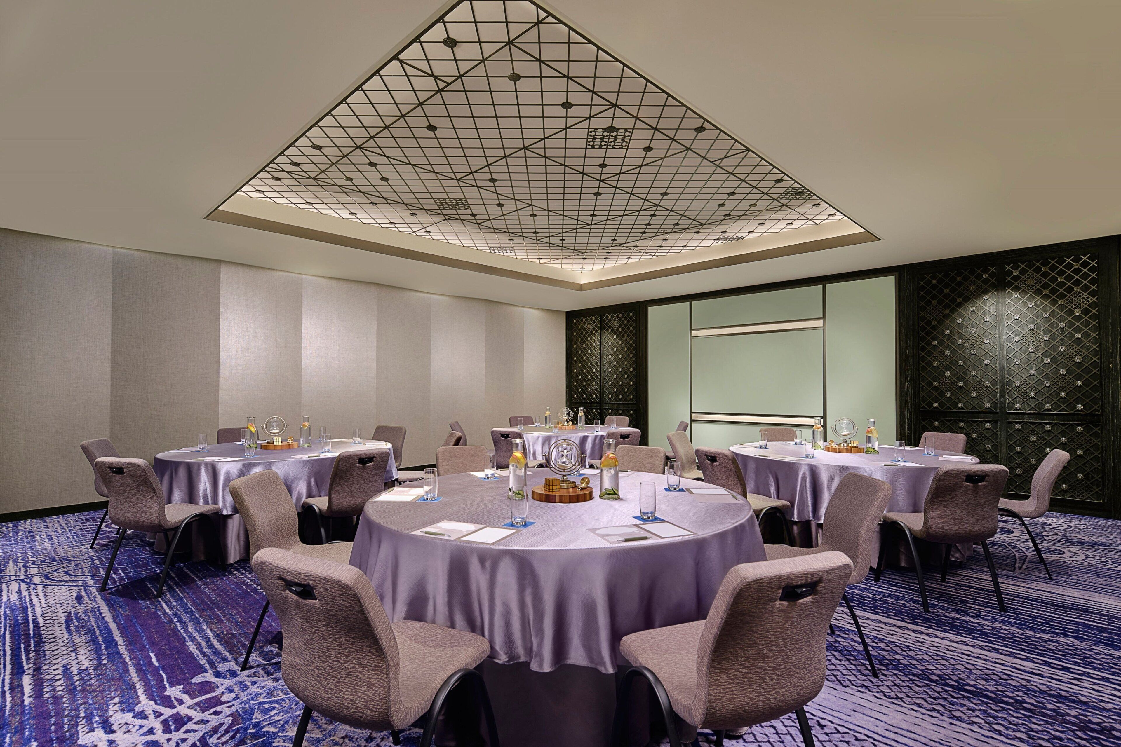Un salón de conferencias con mesas redondas, sillas y un techo con diseño geométrico.