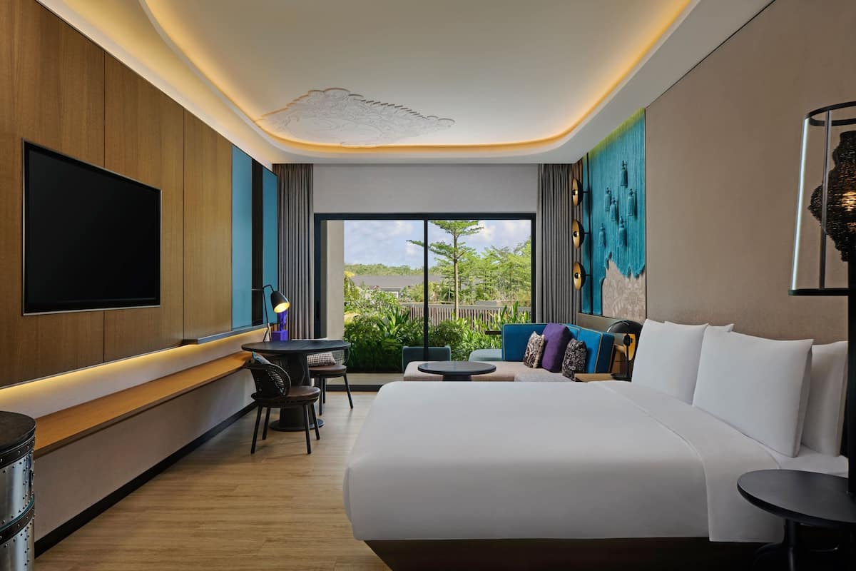 Una habitación de hotel moderna con una cama grande, un televisor de pantalla plana, una pequeña mesa de comedor y un balcón con vistas a zonas verdes.