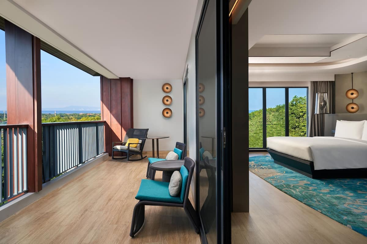 Habitación de hotel moderna con un balcón grande, una cama, zona de estar con sillas y una mesa, y vistas a zonas verdes y paisaje distante.