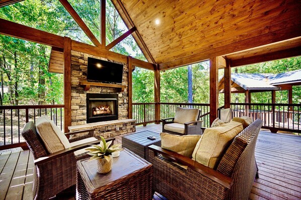 A covered outdoor seating area with a stone fireplace, wicker furniture, and a view of the surrounding forest.