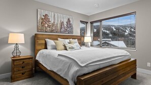 A bedroom with a wooden bed, two bedside tables with lamps, a large window with a view of a snowy landscape, and a wall-mounted artwork.