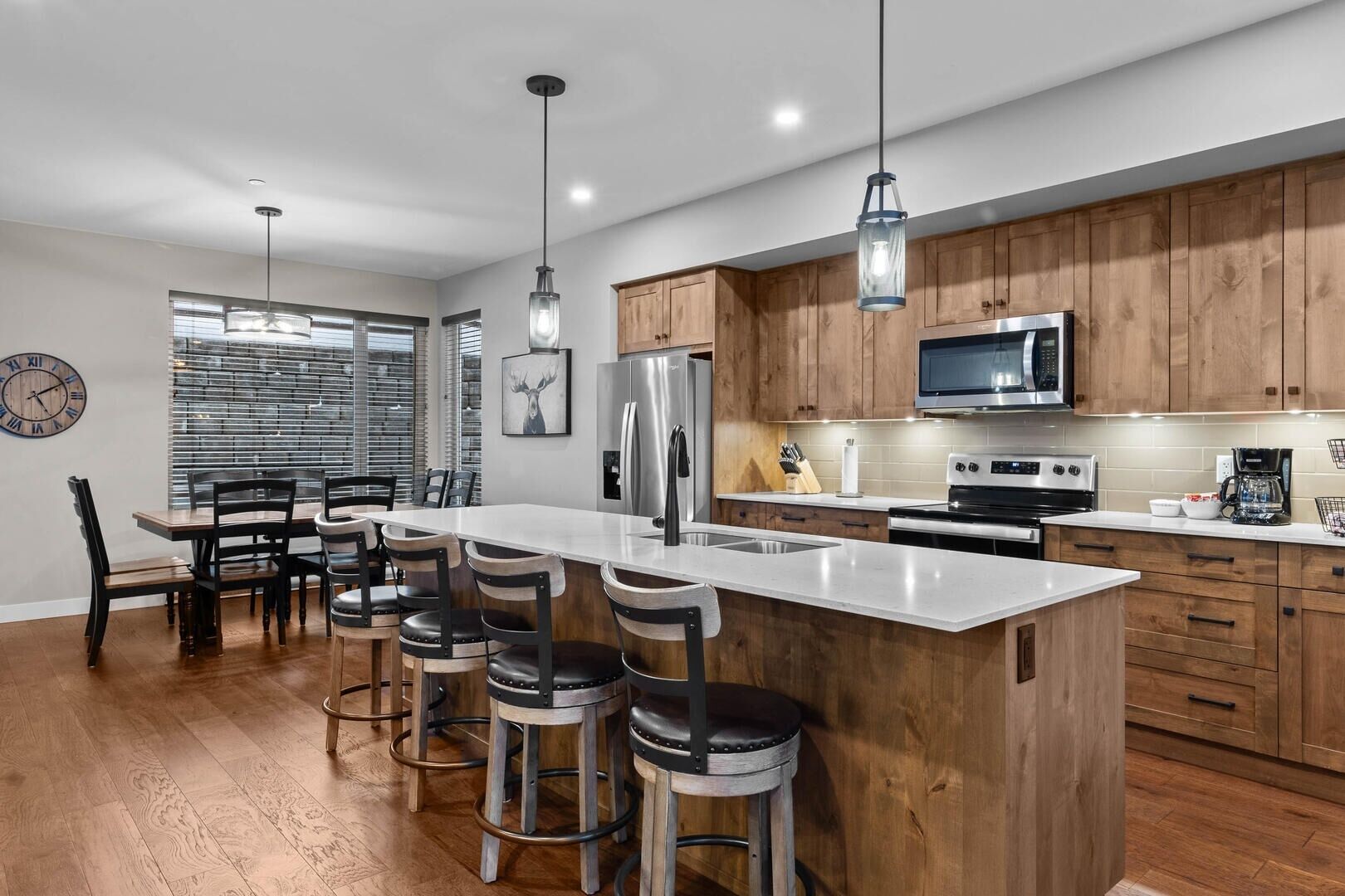 A modern kitchen with wooden cabinets, stainless steel appliances, a large island with bar stools, and a dining area with a clock and blinds.