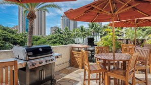 An outdoor patio with a grill, wooden chairs, and a table under a large umbrella, overlooking a cityscape with tall buildings and palm trees. - *Luana Waikiki*No Resort Fees* (Honolulu)
