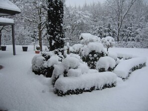 A snow-covered garden with a gazebo, potted plants, and a tall evergreen tree.