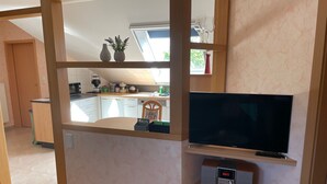 Un salon moderne doté d’un téléviseur à écran plat, d’étagères en bois, d’une petite table et offrant une vue sur la cuisine grâce à une cloison en verre.