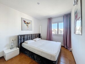 A bedroom with a large bed, a nightstand, a wall-mounted lamp, a window with curtains, and a cityscape view.