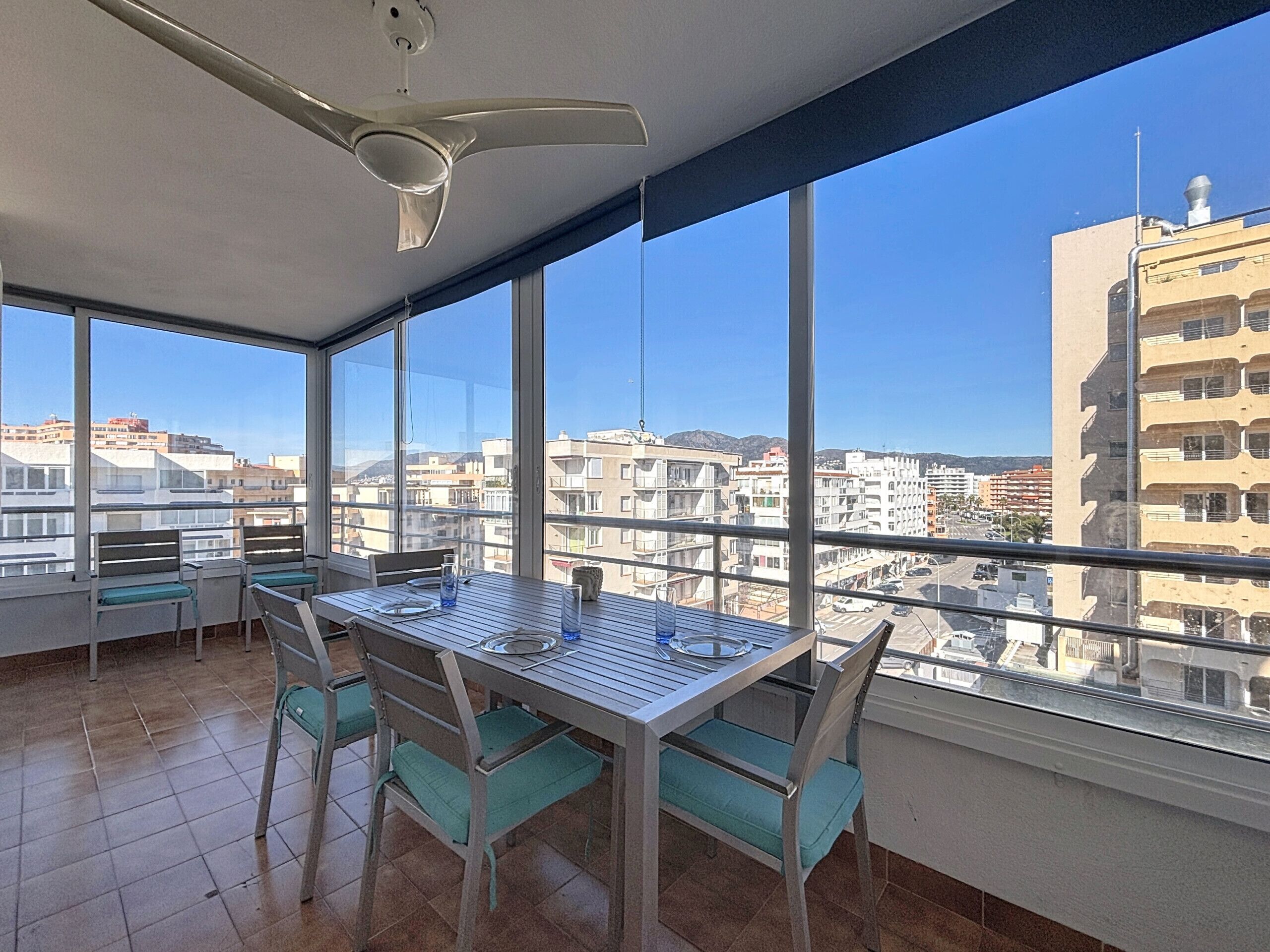 A balcony with a table and chairs, large windows offering a city view, and a ceiling fan.