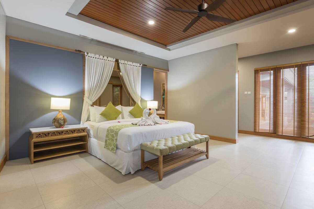 A bedroom with a bed, bedside tables, a bench, a ceiling fan, and a view of a wooden door.