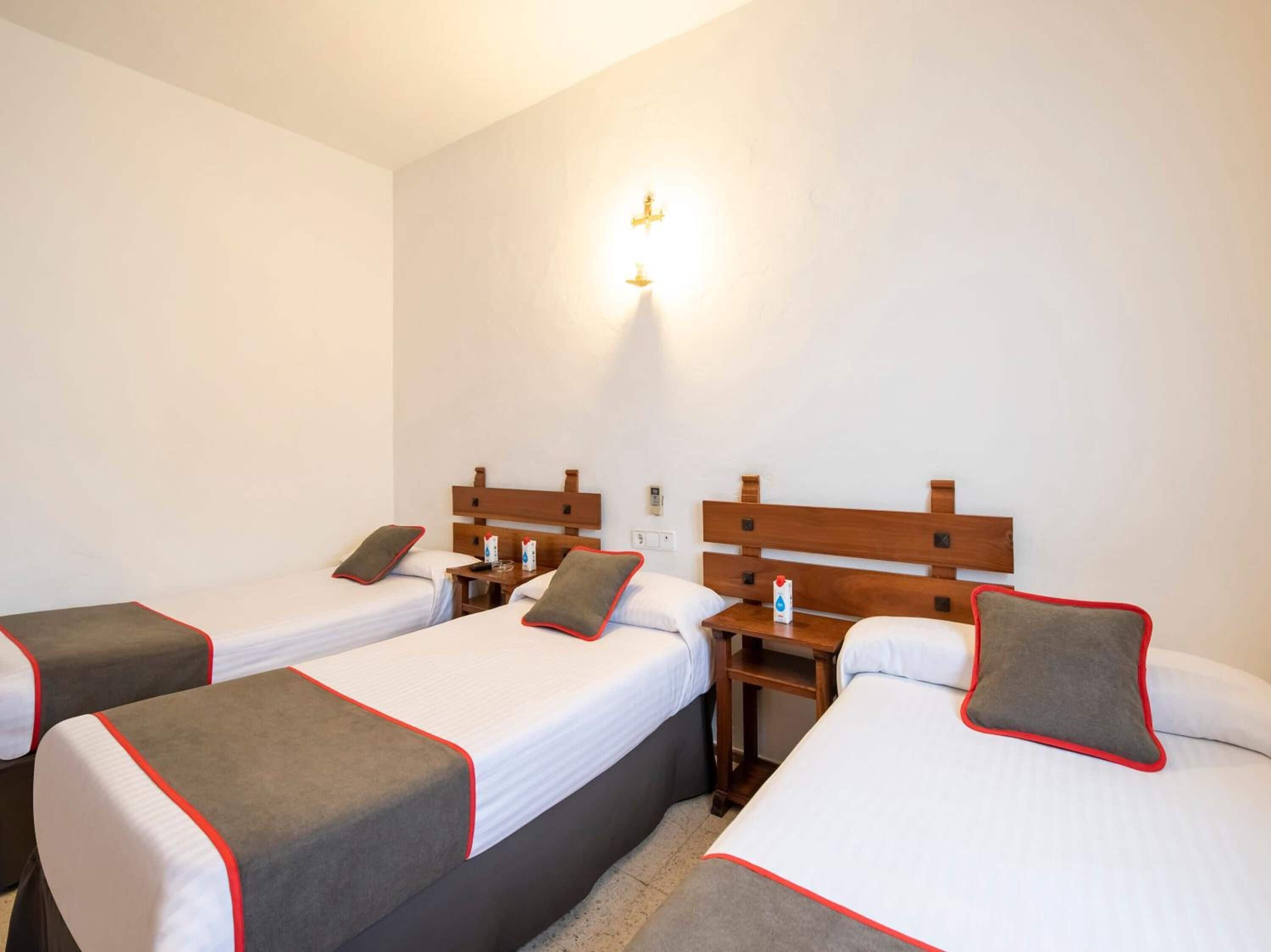 Une chambre d’hôtel avec trois lits simples, chacun doté d’une tête de lit en bois et d’une touche de couleur rouge et grise. Chaque lit est agrémenté d’une table de chevet sur laquelle se trouvent une bouteille et un verre.