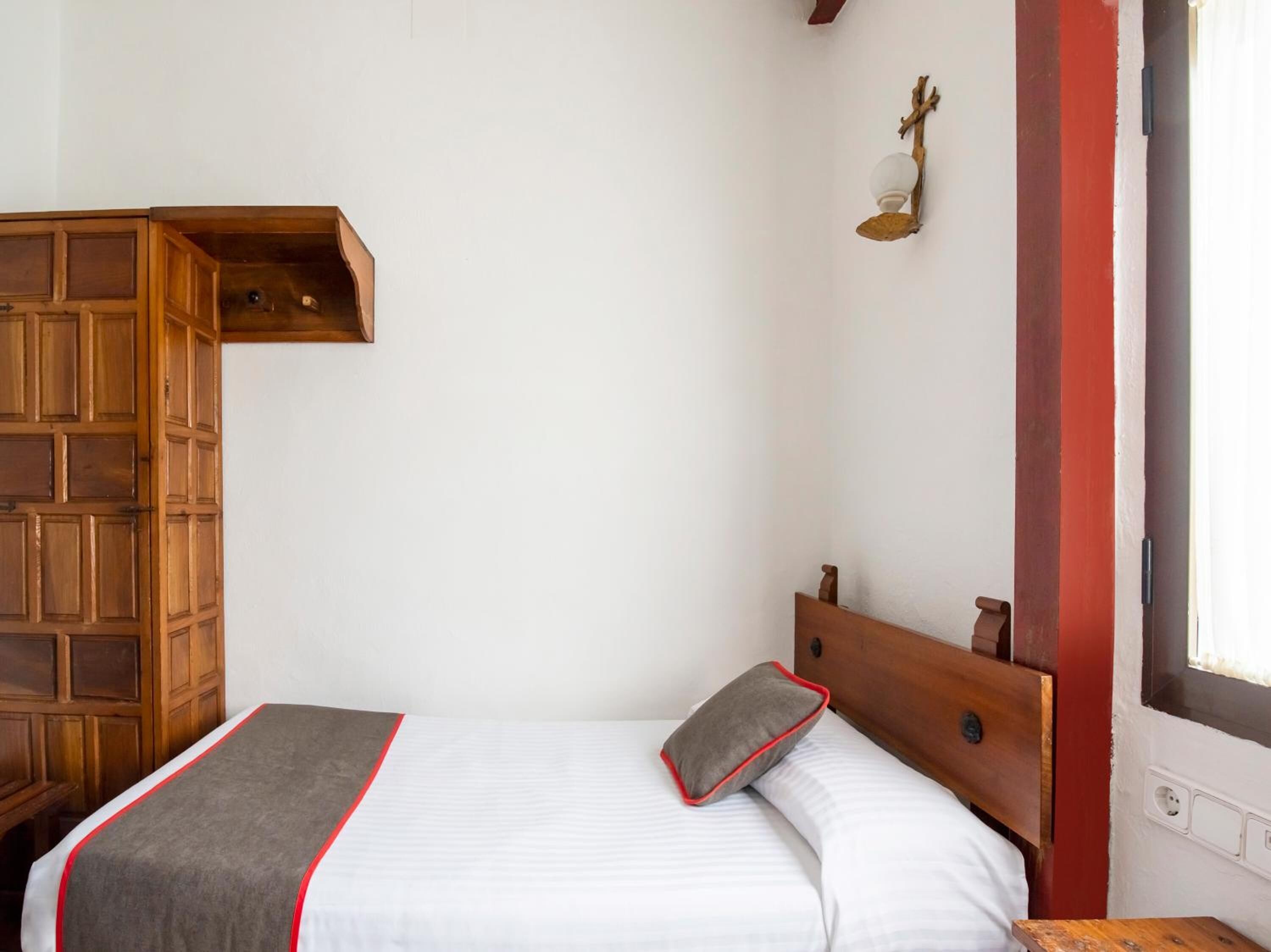 A bedroom with a wooden headboard, a bed with white bedding and a grey pillow, a wooden shelf unit, and a wall-mounted light fixture.