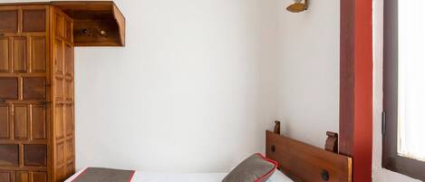 A bedroom with a wooden headboard, a bed with white bedding and a grey pillow, a wooden shelf unit, and a wall-mounted light fixture.
