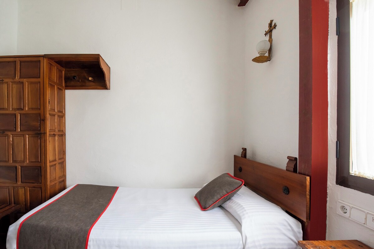 Una cama individual con cabecera de madera, una mesita de noche, una lámpara de pared y un estante de madera.