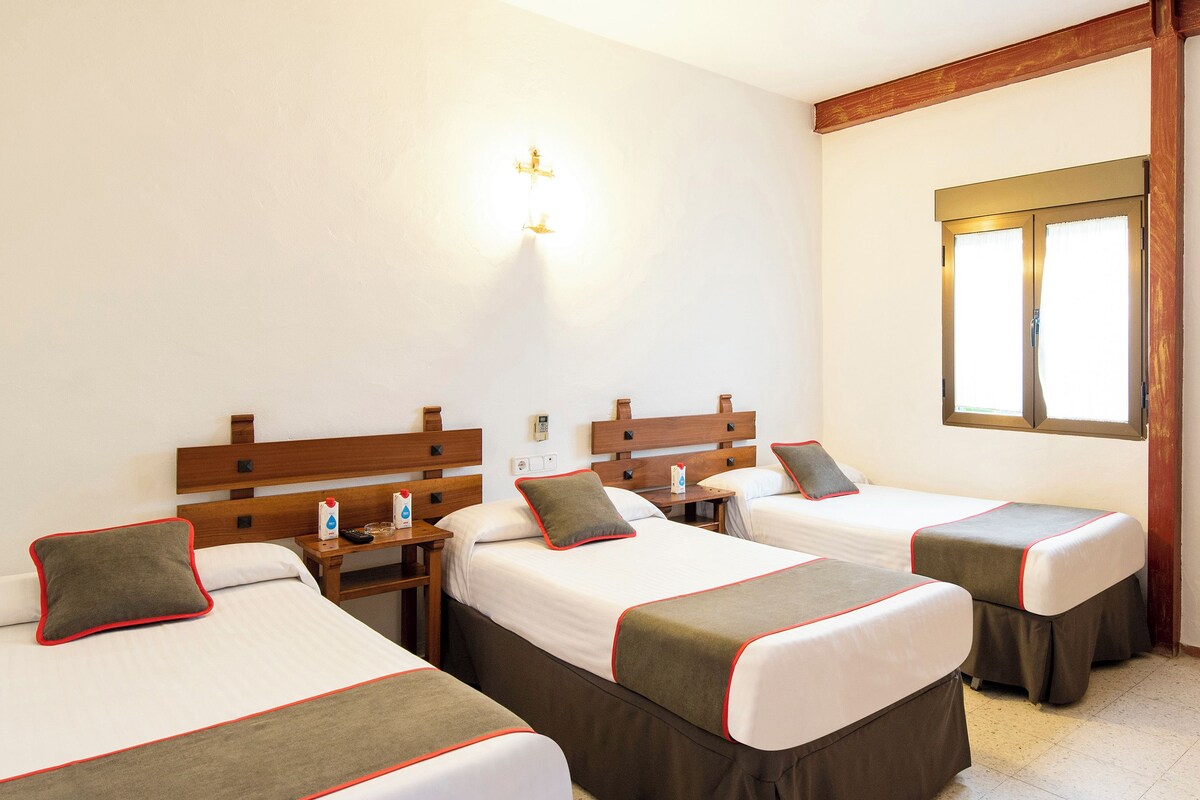 Una habitación de hotel con tres camas individuales, cabeceros de madera, mesitas de noche y una ventana con cortinas.