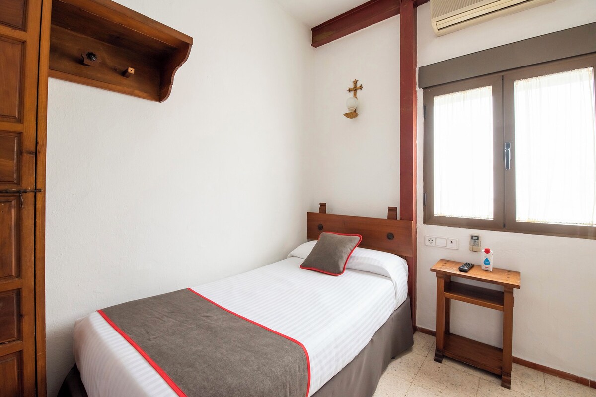 Una cama bien tendida con edredón gris y blanco, una mesita de noche con una botella, y una ventana con cortinas.