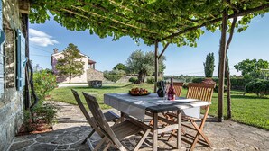 An outdoor dining area with a table and chairs under a pergola, surrounded by stone flooring and greenery. - Rustic Villa Lavanda - Pool & Garden (Koper)