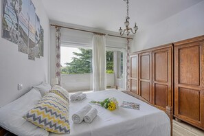 A bedroom with a bed, curtains, a chandelier, a wooden wardrobe, and a window with a view.