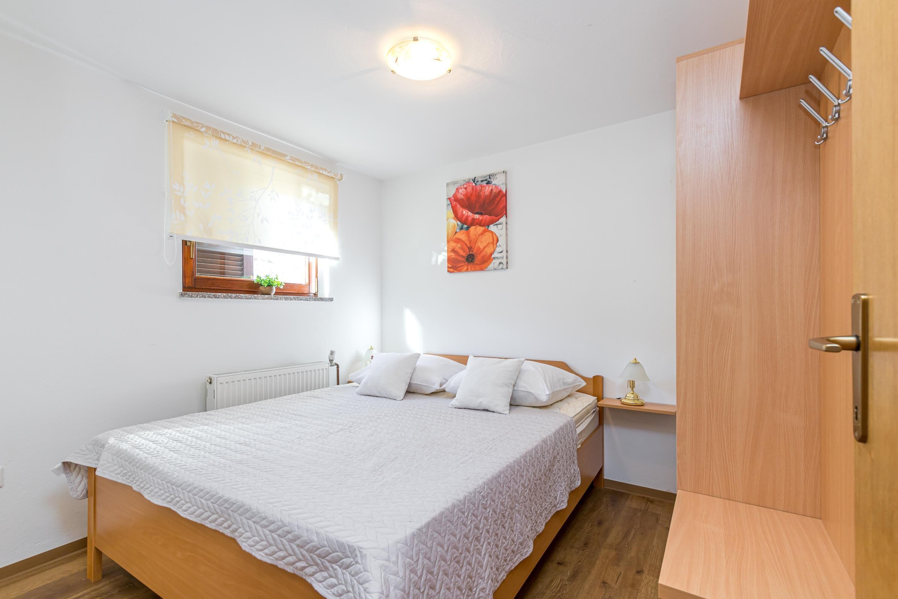Une chambre avec un lit en bois, du linge de lit blanc, un radiateur, une fenêtre avec des stores, une œuvre d’art accrochée au mur et une armoire en bois.
