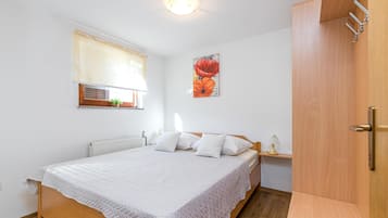 A bedroom with a wooden bed, white bedding, a radiator, a window with blinds, a wall-mounted artwork, and a wooden wardrobe.