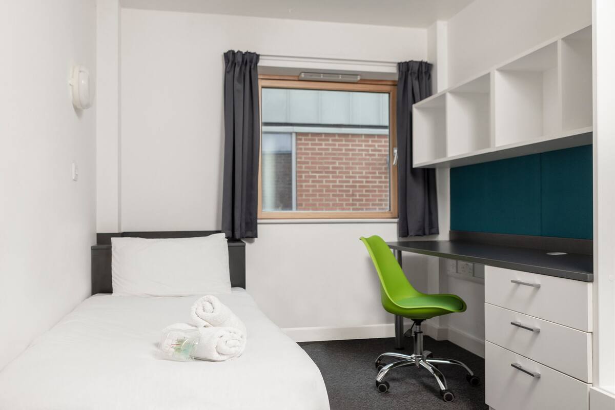 Quarto de hotel com cama, escrivaninha, uma cadeira verde e janela com cortinas.