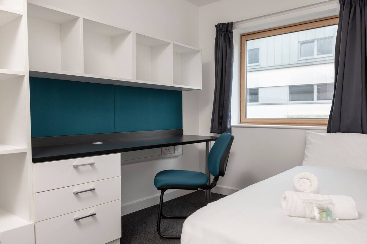 Quarto de hotel com cama, escrivaninha com cadeira, janela com cortinas e vista para um edifício.