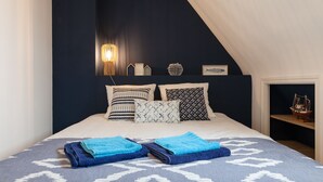 Ein Schlafzimmer mit dunkelblauer Akzentwand, geneigter Decke und einem Bett mit gemusterten Textilien und vielen Kissen.